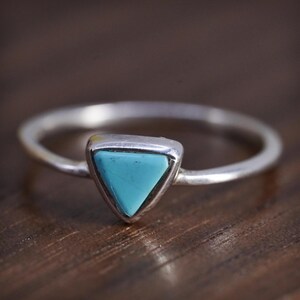 May include: A silver ring featuring a small, triangular-shaped turquoise stone set in a smooth metal bezel. The band is thin and polished, showcasing a simple design that highlights the bright blue color of the stone centered on the ring.