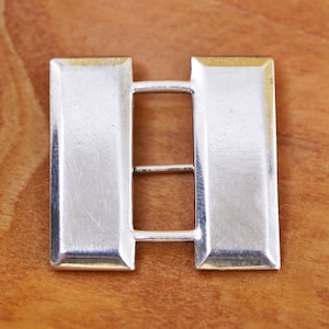 Peut inclure: Insigne de grade militaire en métal argenté, composé de deux barres rectangulaires reliées par deux petites barres. L'insigne est posé sur une surface en bois, mettant en valeur sa surface polie et réfléchissante.