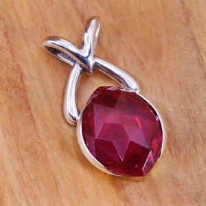 May include: A silver pendant with a large, faceted, oval-shaped, deep red gemstone. The pendant has a loop at the top and a curved design. The gemstone is set in a silver bezel.