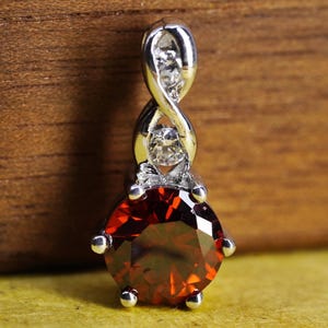 May include: A silver-toned pendant featuring a large, round, faceted deep red gemstone at the base. Above the main stone, the metal forms a twisted, infinity-style loop design that is accented with two small, clear, round, faceted stones.