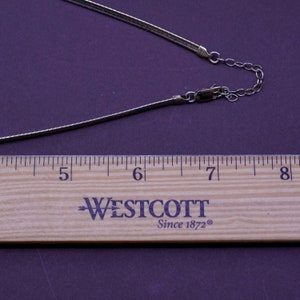 May include: A silver-toned necklace with a slender, flexible chain. The necklace is shown with a wooden ruler, measuring approximately 5 to 8 inches. The background is a solid, dark purple.