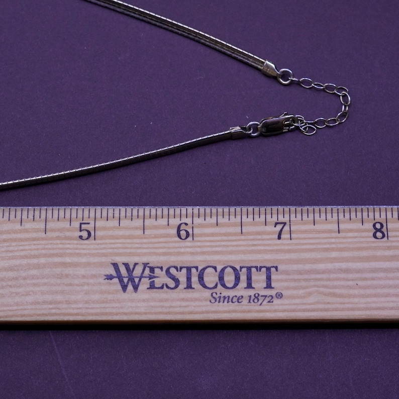 May include: A silver-toned necklace with a slender, flexible chain. The necklace is shown with a wooden ruler, measuring approximately 5 to 8 inches. The background is a solid, dark purple.