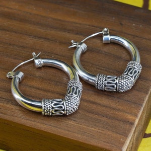 May include: A pair of silver hoop earrings with decorative detailing. The earrings have a thick, rounded design with textured patterns on the lower portion. The metal is shiny and reflective, and the earrings are displayed on a wooden surface.