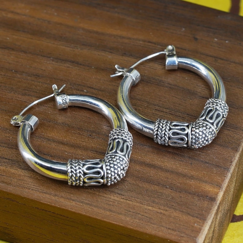 May include: A pair of silver hoop earrings with decorative detailing. The earrings have a thick, rounded design with textured patterns on the lower portion. The metal is shiny and reflective, and the earrings are displayed on a wooden surface.