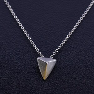 May include: A silver necklace featuring a geometric triangular pendant. The pendant has a brushed metal finish with a slight gold tone at the bottom. The chain is a delicate link style, suitable for everyday wear.