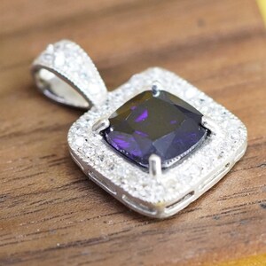 May include: A silver pendant with a square, deep purple gemstone at the center, surrounded by a halo of smaller, clear stones. The pendant has a decorative bail for a chain and is set against a wooden surface.