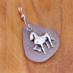 May include: A silver-colored horse pendant on a lavender-colored, teardrop-shaped stone. The pendant is attached to a silver chain. The background is a light brown wooden surface. The horse is in a trotting pose.