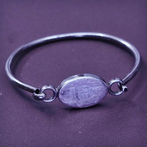 May include: A silver bangle bracelet with an oval centerpiece. The centerpiece has engraved initials. The bracelet has a simple design with a clasp. The bracelet is approximately 17.8 cm in circumference.