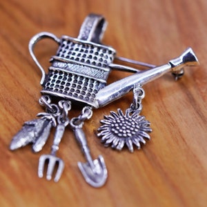 Puede incluir: Broche plateado con forma de regadera, con colgantes de un girasol, una pequeña pala, un tenedor y una hoja. El broche se presenta sobre un fondo de madera cálida.