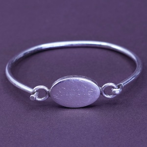 May include: A silver bangle bracelet with an oval centrepiece. The centrepiece has the engraved word "Stadie". The bracelet is a simple, elegant design, perfect for everyday wear. The bracelet is made of a shiny, silver metal.