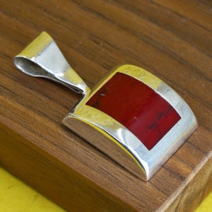 May include: A silver pendant with a rectangular, red stone inlay. The stone is set within a silver frame, and the pendant has a curved top for a chain. The pendant is displayed on a wooden surface.
