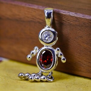 Peut inclure: Pendentif argenté en forme de figurine stylisée. Le pendentif présente une pierre précieuse claire et ronde en haut et une grande pierre précieuse ovale rouge foncé au centre. De petites perles rondes en argent forment les bras et les pieds.