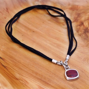 May include: A black cord necklace with a silver pendant. The pendant has a square, reddish-brown stone set in a silver frame. The necklace is displayed on a light brown wooden surface.