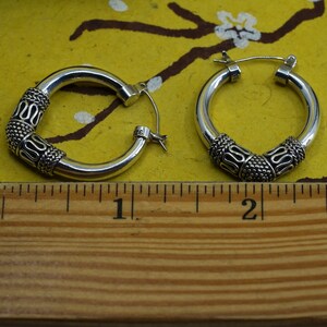 May include: A pair of silver hoop earrings. Each earring has a decorative band with an intricate pattern. The earrings are shown against a yellow background with a ruler for scale.