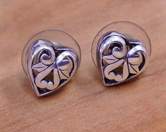 May include: A pair of silver-toned heart-shaped stud earrings. Each earring features an openwork design with swirling patterns and a central leaf motif. The earrings are set against a light brown wooden surface.
