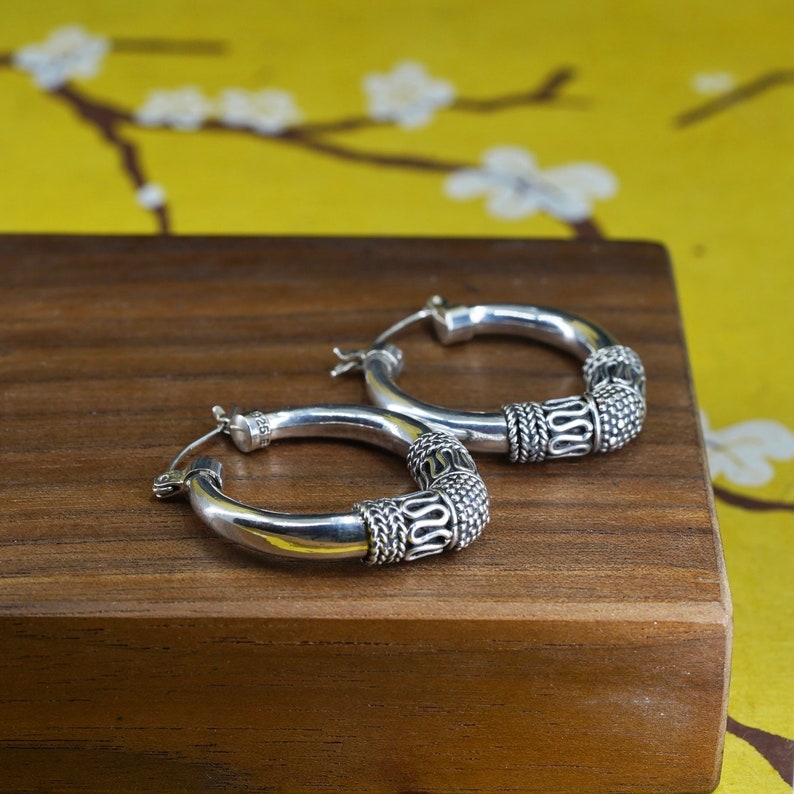 May include: A pair of silver hoop earrings with decorative detailing. The earrings have a polished finish and feature a textured design on the lower portion of the hoops. The earrings are displayed on a wooden surface against a yellow background.