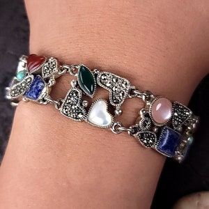 Könnte beinhalten: Ein silbernes Armband mit herzförmigem Design zeigt eine Vielzahl von bunten Edelsteinen, darunter Rot, Grün, Blau und Weiß.