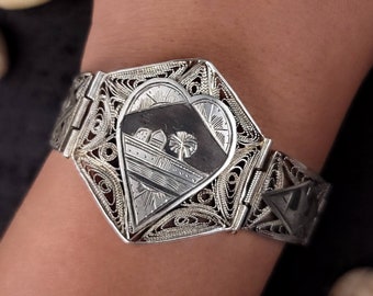 Puede incluir: Un brazalete de filigrana plateado con un colgante en forma de corazón que presenta un diseño grabado en blanco y negro de una casa y árboles.