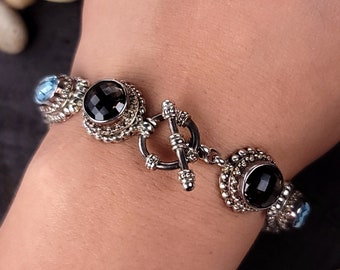 Puede incluir: Un brazalete de plata con cierre de palanca presenta piedras preciosas negras y azules. El brazalete está hecho de plata de ley y tiene un diseño de cuentas.