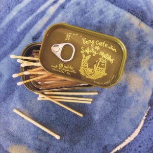 Puede incluir: Una lata verde oliva abierta de cerillas de seguridad con el texto "Very Cute Tin of Safety Matches" e ilustraciones. La lata está sobre un plato de cerámica azul, con varias cerillas esparcidas.