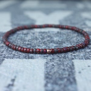 Könnte beinhalten: Ein Armband aus Perlen in Dunkelrot und Grau. Das Armband besteht aus kleinen, runden Perlen, mit einer einzelnen kupferfarbenen Perle. Das Armband ist für das Tragen am Handgelenk konzipiert.