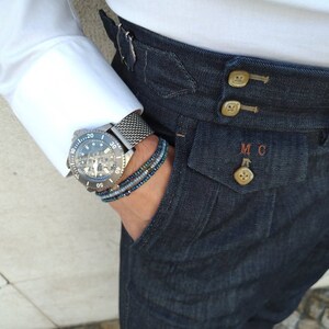 May include: A close-up of a person wearing a white shirt and dark blue jeans with a silver watch and a beaded bracelet on their wrist. The jeans have a button fly and a pocket with the initials "MC" embroidered on it.