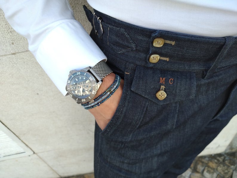 May include: A close-up of a person wearing a white shirt and dark blue jeans with a silver watch and a beaded bracelet on their wrist. The jeans have a button fly and a pocket with the initials "MC" embroidered on it.