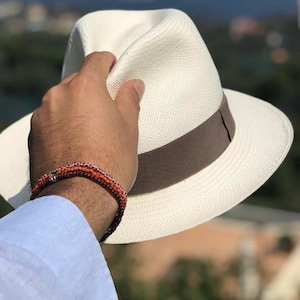 May include: A white Panama hat with a brown band. The hat is being held by a hand wearing a multi-colored beaded bracelet.