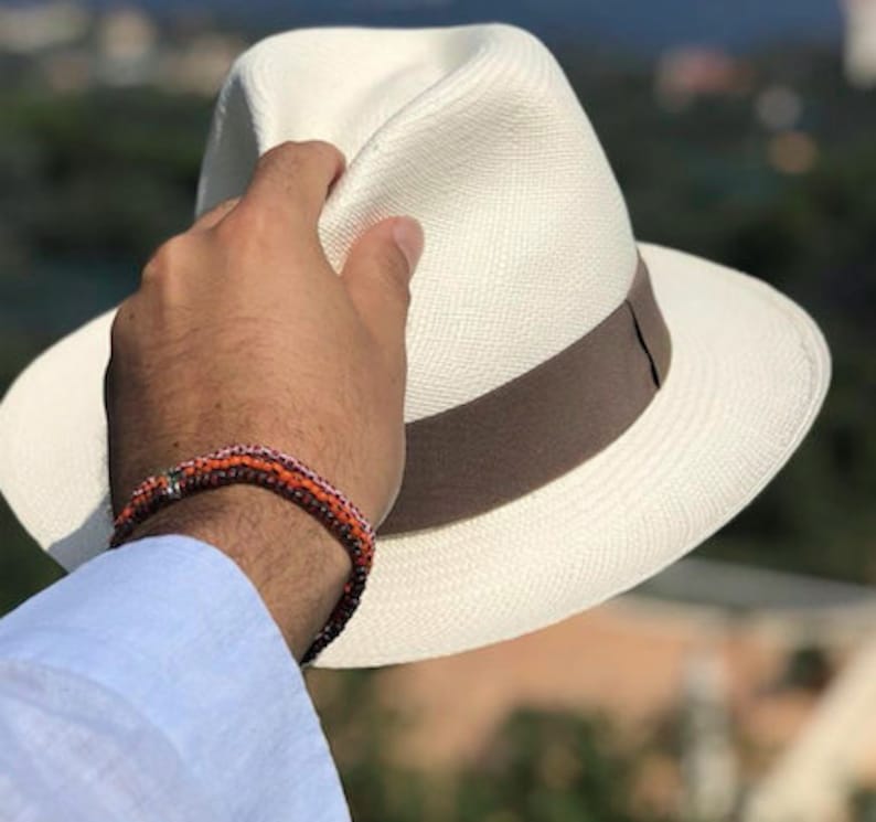 May include: A white Panama hat with a brown band. The hat is being held by a hand wearing a multi-colored beaded bracelet.