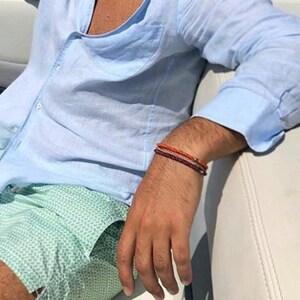 May include: A man wearing a light blue linen shirt and green patterned swim trunks. He is wearing a stack of bracelets on his wrist.