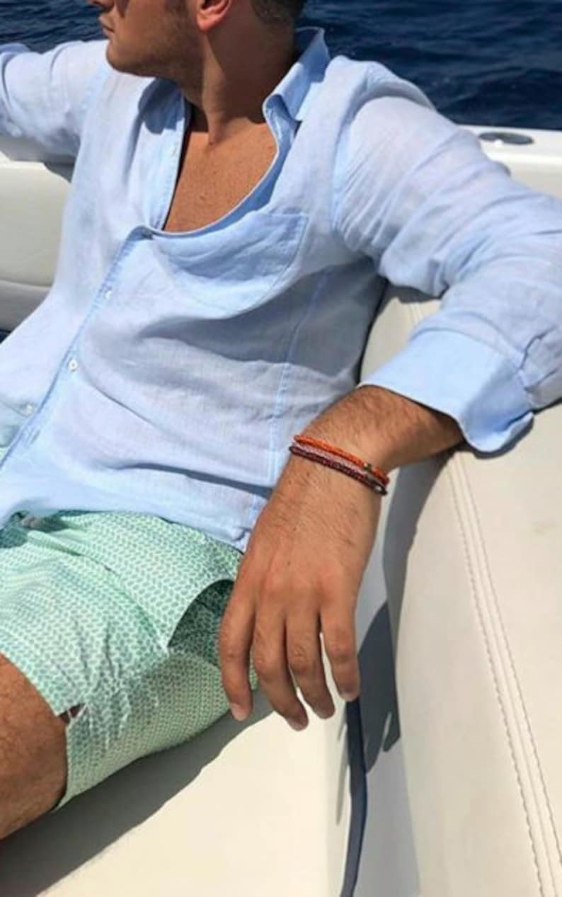 May include: A man wearing a light blue linen shirt and green patterned swim trunks. He is wearing a stack of bracelets on his wrist.