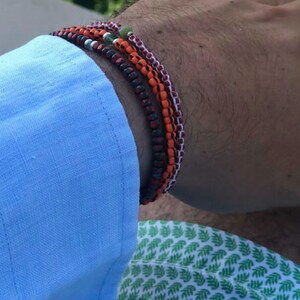 May include: A stack of four bracelets on a person's wrist. The bracelets are made of small beads in shades of red, orange, and white. One bracelet has a green bead.