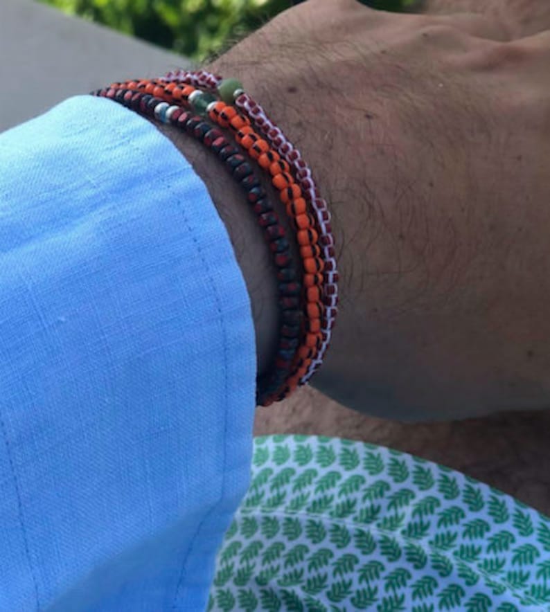May include: A stack of four bracelets on a person's wrist. The bracelets are made of small beads in shades of red, orange, and white. One bracelet has a green bead.