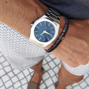 May include: A silver watch with a blue face and a black and blue beaded bracelet. The watch has a square face with rounded corners and the text "ST. MILAN" on the face. The watch is on a person's wrist.