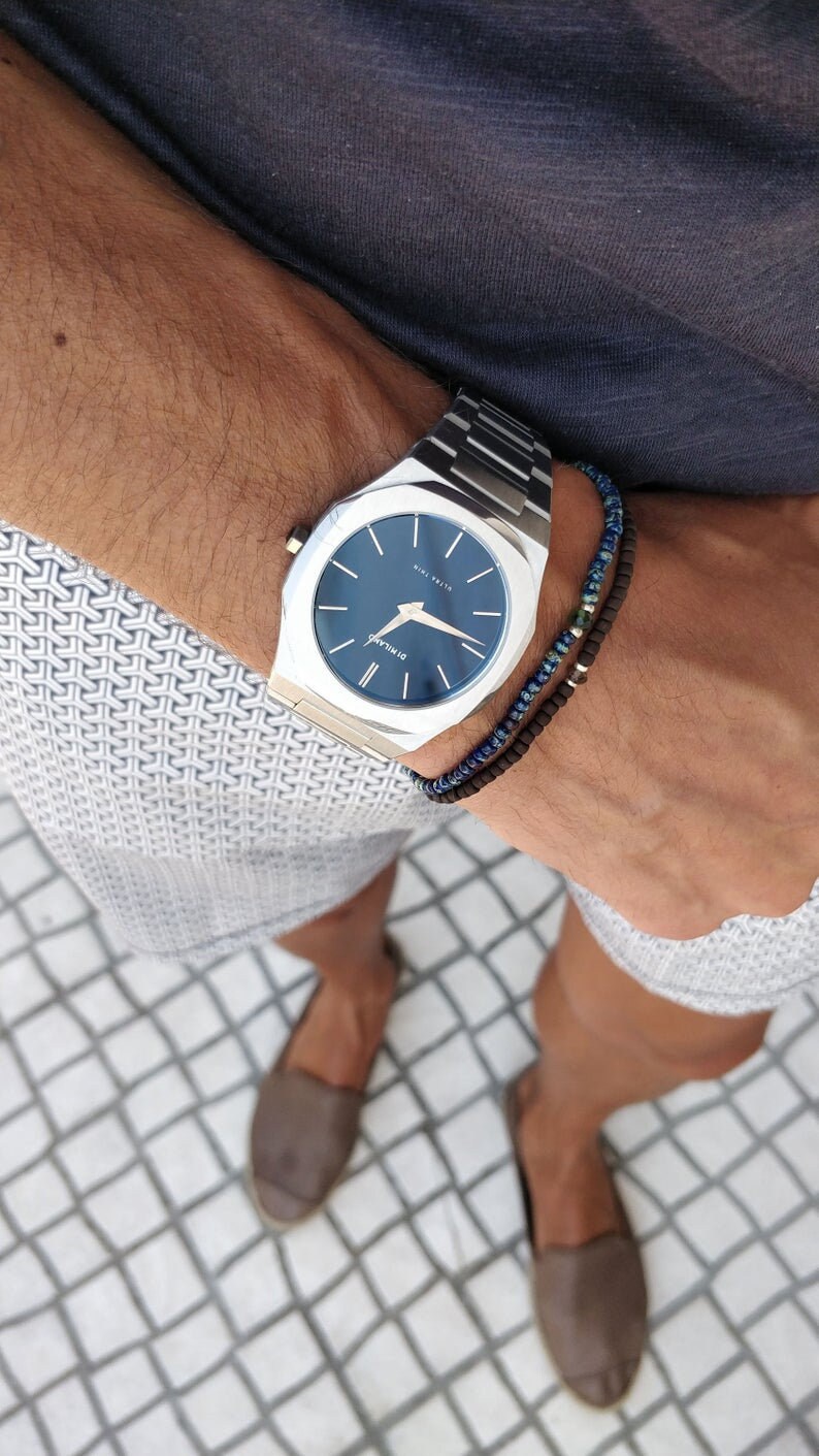 May include: A silver watch with a blue face and a black and blue beaded bracelet. The watch has a square face with rounded corners and the text "ST. MILAN" on the face. The watch is on a person's wrist.