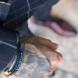 Puede incluir: Una mu&ntilde;eca de hombre con un tatuaje de calavera y dos pulseras de cuentas, una azul y otra marr&oacute;n, con una pulsera de oro en la parte superior.