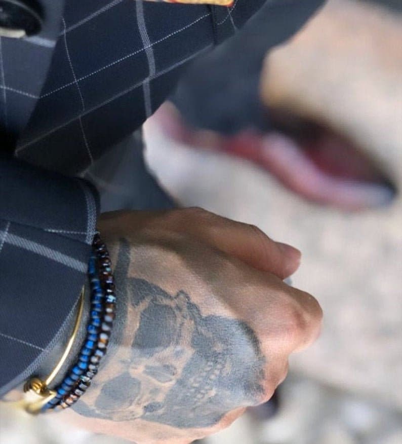 Puede incluir: Una mu&ntilde;eca de hombre con un tatuaje de calavera y dos pulseras de cuentas, una azul y otra marr&oacute;n, con una pulsera de oro en la parte superior.