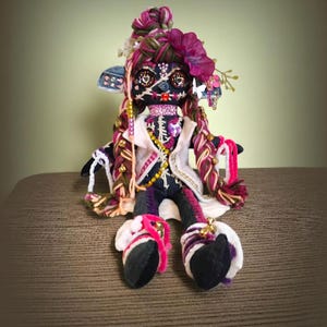 May include: A handmade doll with a Day of the Dead theme. The doll has a black face with painted details, braids, and a floral headpiece. It wears a white jacket, a beaded necklace, and colorful clothing. The doll is seated on a brown surface.