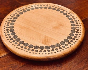 May include: A round wooden serving board with a decorative border of dark gray dots. The board is made of light-colored wood and has a smooth surface. The dots are arranged in a circular pattern around the edge of the board.