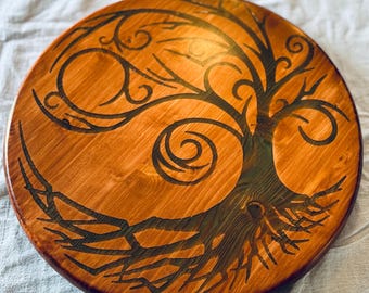 May include: A round wooden decorative piece with a tree of life design. The tree is depicted in black against a warm, reddish-brown wood grain background. The tree's branches and roots are intricately detailed.