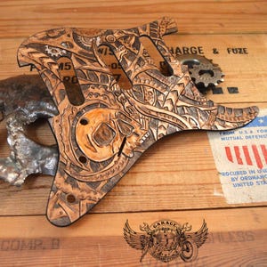 May include: A brown and tan laser-cut guitar pickguard featuring a skull and gear design. The pickguard is displayed on a wooden surface alongside a metal skull and a gear. The image also includes text and a logo.