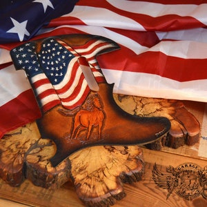 May include: A leather boot shaped wall decoration featuring an American flag and a horse design. The boot is mounted on a wooden base and surrounded by an American flag.