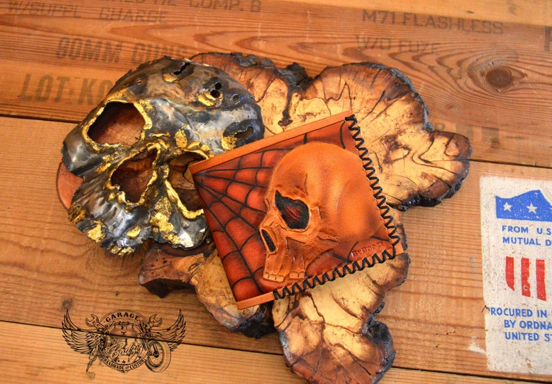 May include: A brown leather wallet with a skull and spiderweb design. The wallet is stitched with a black thread and has a zipper closure.
