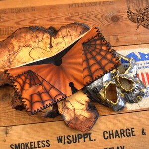 May include: A brown leather wallet with a spiderweb design and a metal skull mask. The wallet is on a wooden surface with text that reads "M71 FLASHLESS" and "SMOKELESS W/SUPPL. CHARGE & FUZE".