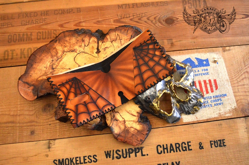 May include: A brown leather wallet with a spiderweb design and a metal skull mask. The wallet is on a wooden surface with text that reads "M71 FLASHLESS" and "SMOKELESS W/SUPPL. CHARGE & FUZE".