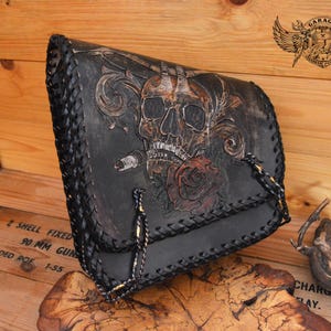 Pode incluir: Bolsa de couro preta com design de caveira, rosa e charuto. A bolsa tem uma borda trançada e alças de corrente douradas. A imagem também mostra o texto "GARAGE".