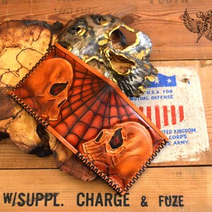 May include: A brown leather wallet with a spiderweb design and two skull motifs. The wallet has a stitched edge and is displayed on a wooden surface.