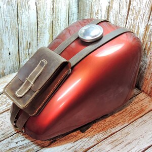 May include: A brown leather motorcycle tank bag with a strap attached to a red gas tank. The bag has a flap with a buckle closure.