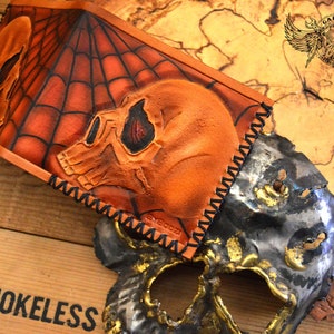 May include: A brown leather wallet with a spiderweb design and two skull motifs. The wallet has black stitching around the edges.