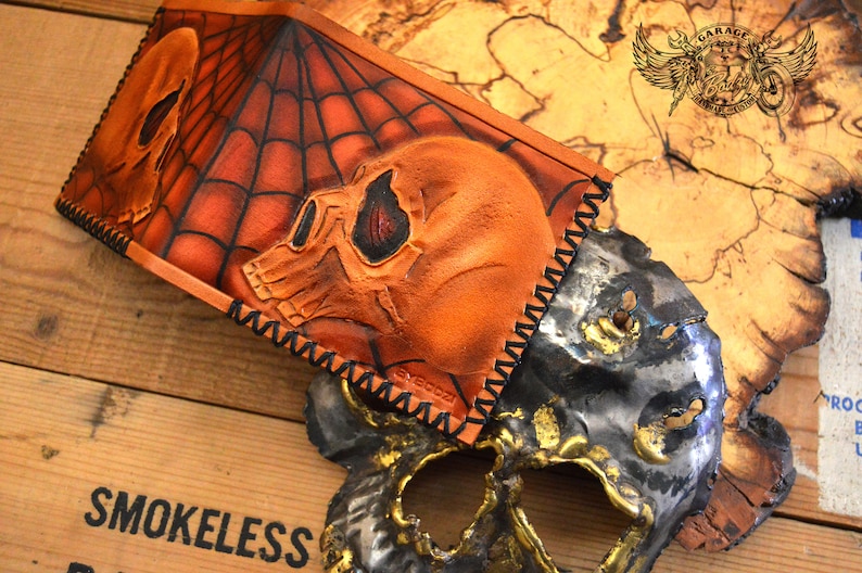 May include: A brown leather wallet with a spiderweb design and two skull motifs. The wallet has black stitching around the edges.